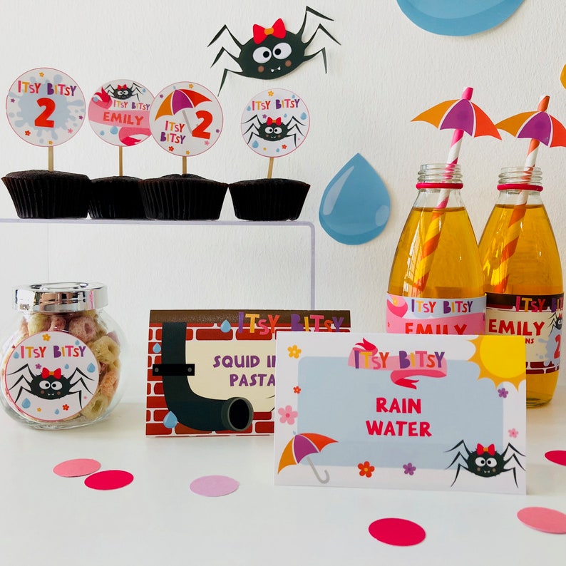 May include: A table setting for a birthday party with a Itsy Bitsy theme. There are cupcakes with spider web toppers, a jar of cereal, and bottles of juice with labels that say "Itsy Bitsy Emily". There are also signs that say "Squid Ink Pasta" and "Rain Water".