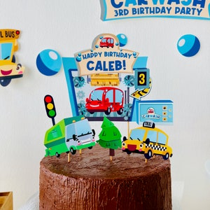 A chocolate cake decorated with a car wash themed cake topper. The topper features a red car, a green refuse truck, a yellow taxi, a green tree, and a sign that says "Happy Birthday Caleb!" with the number 3.