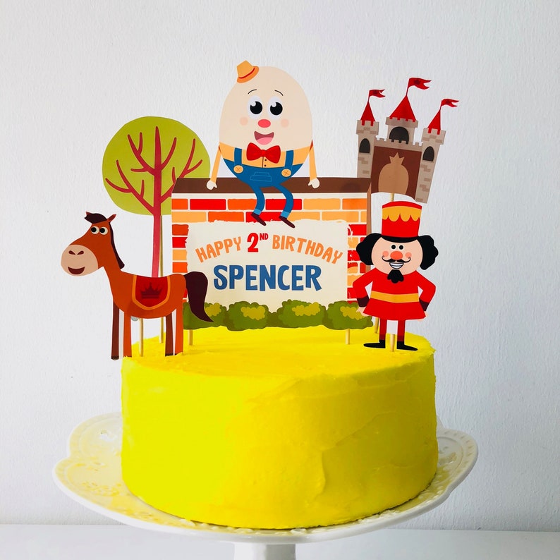 Puede incluir: Un pastel amarillo decorado con una pared de ladrillo, un caballo, un hombre de jengibre, un castillo y un personaje de huevo en la parte superior. El pastel est&aacute; decorado con el texto "Happy 2nd Birthday Spencer".