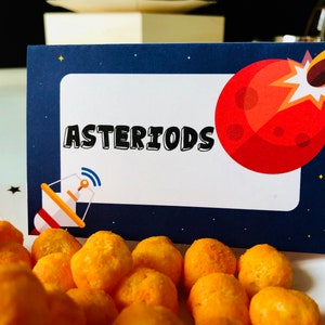 May include: A navy blue card with the word "ASTEROIDS" in black and white. A red planet with a flame graphic is on the right. A radar dish graphic is on the left. Orange cheese puffs are in the foreground.