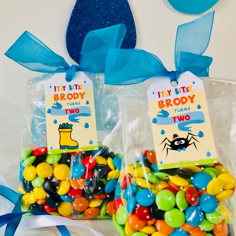 May include: Two clear plastic bags filled with colorful candy, each with a tag attached. The tags are yellow with blue and red accents. The text on the tags reads "Itsy Bitsy Brody Turns Two". One tag features a cartoon spider and the other a cartoon boot.