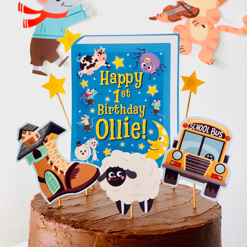 May include: A chocolate cake with a blue book topper that says "Happy 1st Birthday Ollie!" and other toppers including a boot, a sheep, and a school bus.