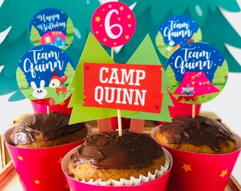 May include: A set of six cupcake toppers with a camping theme. The toppers feature a green and brown tree, a red and orange sign that says "Camp Quinn", a blue and white circle with the text "Team Quinn", a pink circle with the number "6" and a blue and white circle with the text "Team Quinn".