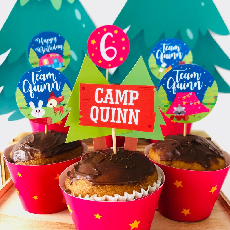 May include: A set of six cupcake toppers with a camping theme. The toppers feature a green and brown tree, a red and orange sign that says "Camp Quinn", a blue and white circle with the text "Team Quinn", a pink circle with the number "6" and a blue and white circle with the text "Team Quinn".