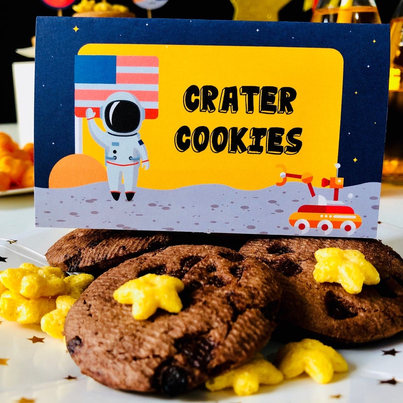 May include: Chocolate chip cookies with star-shaped cereal pieces on a white plate. A sign with the text "CRATER COOKIES" features an astronaut, the American flag, and a space rover against a yellow and blue background. The cookies are a treat for a space-themed party.