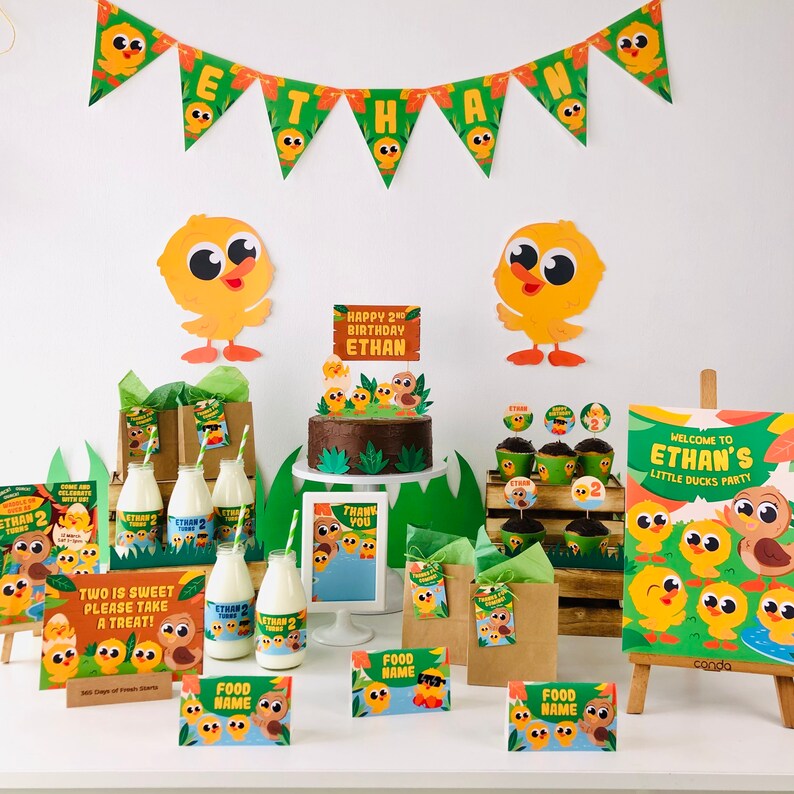 May include: A birthday party table setting with a brown cake decorated with yellow chicks and a sign that says "Happy 2nd Birthday Ethan". There are also yellow chick themed party favors, cupcake toppers, and a sign that says "Welcome to Ethan's Little Ducks Party".