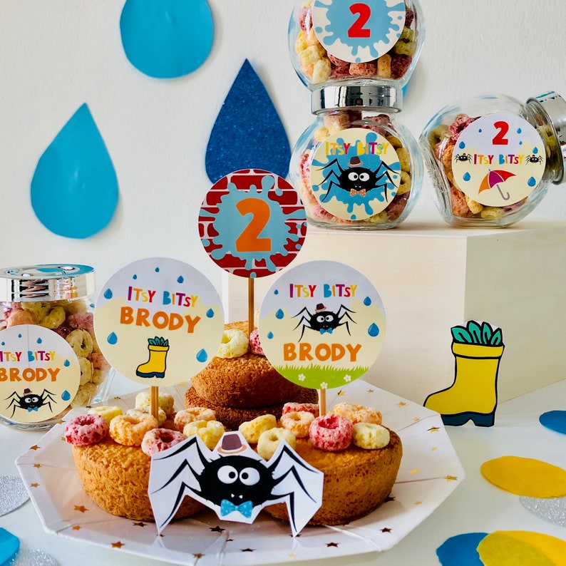 May include: A birthday party centerpiece with a spider, a yellow rain boot, and a cake with sprinkles. The cake is decorated with round paper circles that say "Itsy Bitsy Brody" and "2".