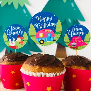 May include: Three cupcakes with chocolate frosting and pink cupcake liners with yellow stars. Each cupcake has a round paper topper with a camping theme. One topper says "Happy Birthday" with a camper van, another says "Team Quinn" with a deer and rabbit, and the last says "Team Quinn" with a tent and raccoon.