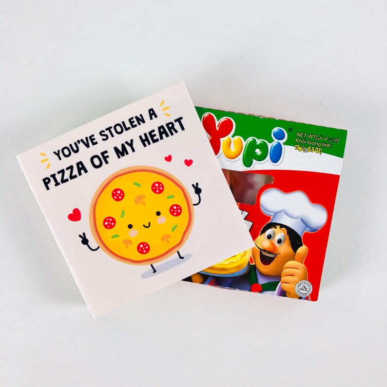 Pu&ograve; includere: Un biglietto bianco con la scritta "YOU'VE STOLEN A PIZZA OF MY HEART" e un'illustrazione di pizza a cartoni animati. Accanto, una scatola di caramelle Yupi con un'illustrazione di chef e la scritta "NET WT 15g (0,5 oz)".
