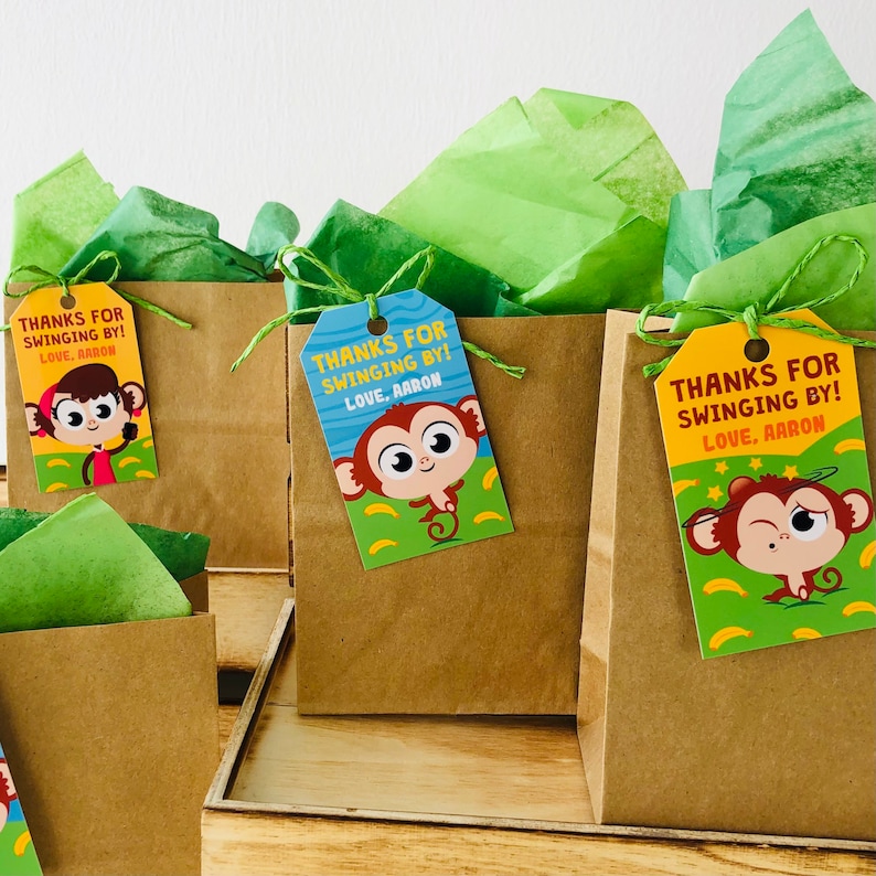 May include: Three brown paper bags with green tissue paper inside. Each bag has a colourful monkey-themed gift tag attached with the text "Thanks for swinging by! Love, Aaron".
