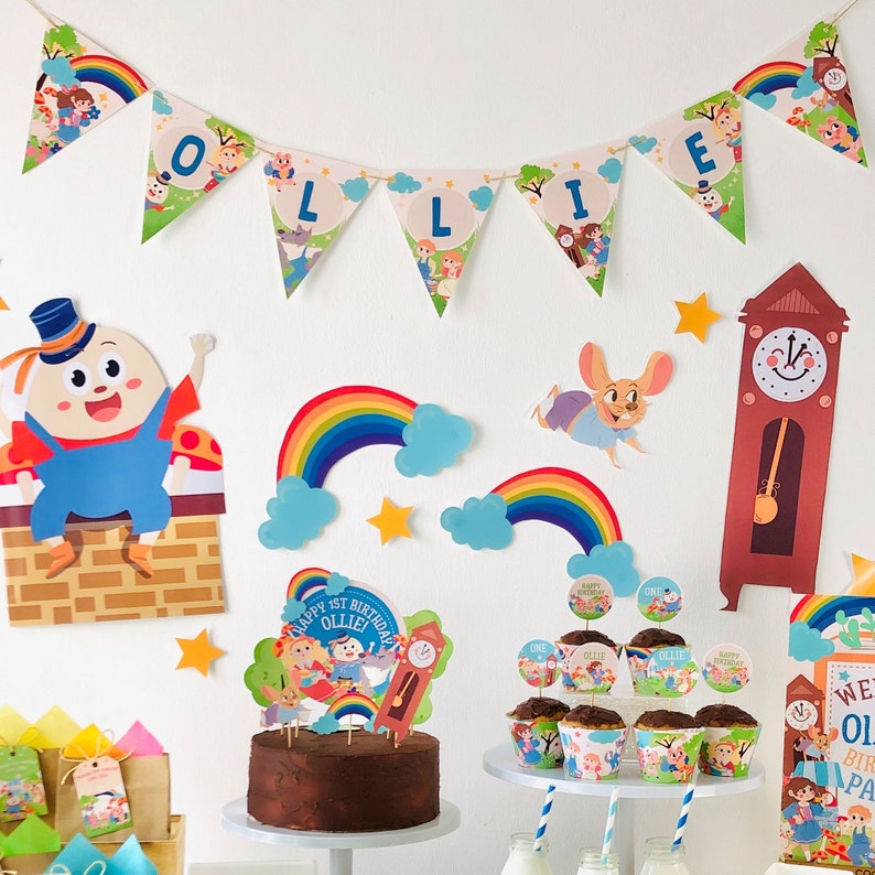 K&ouml;nnte beinhalten: Ein Geburtstags-Setup mit einem 'Happy 1st Birthday Ollie' Kuchentopper, einem Schokoladenkuchen, Cupcakes mit 'One' und 'Ollie' Toppern, einer Uhr, einem Regenbogen und einem Banner mit der Aufschrift 'Ollie'.