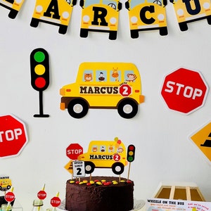 May include: A yellow school bus themed birthday party with decorations including a banner, stop signs, a traffic light, and a cake with a school bus topper. The cake is decorated with yellow and red sweets. The banner says "MARCUS 2".
