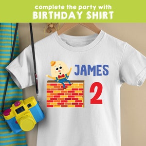 May include: White t-shirt with a cartoon character of a yellow egg with a red bow tie and blue overalls sitting on a brick wall. The text "JAMES 2" is printed on the shirt.