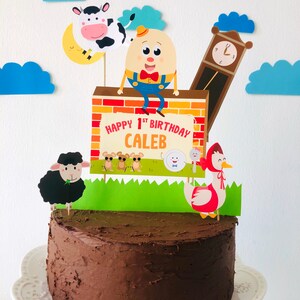 K&ouml;nnte beinhalten: Ein Schokoladenkuchen, der mit einem Banner 'Happy 1st Birthday Caleb', einer Comicfigur eines Eies, einer Kuh, einem Schaf, einer Uhr und einer rotgekappten Gans verziert ist.