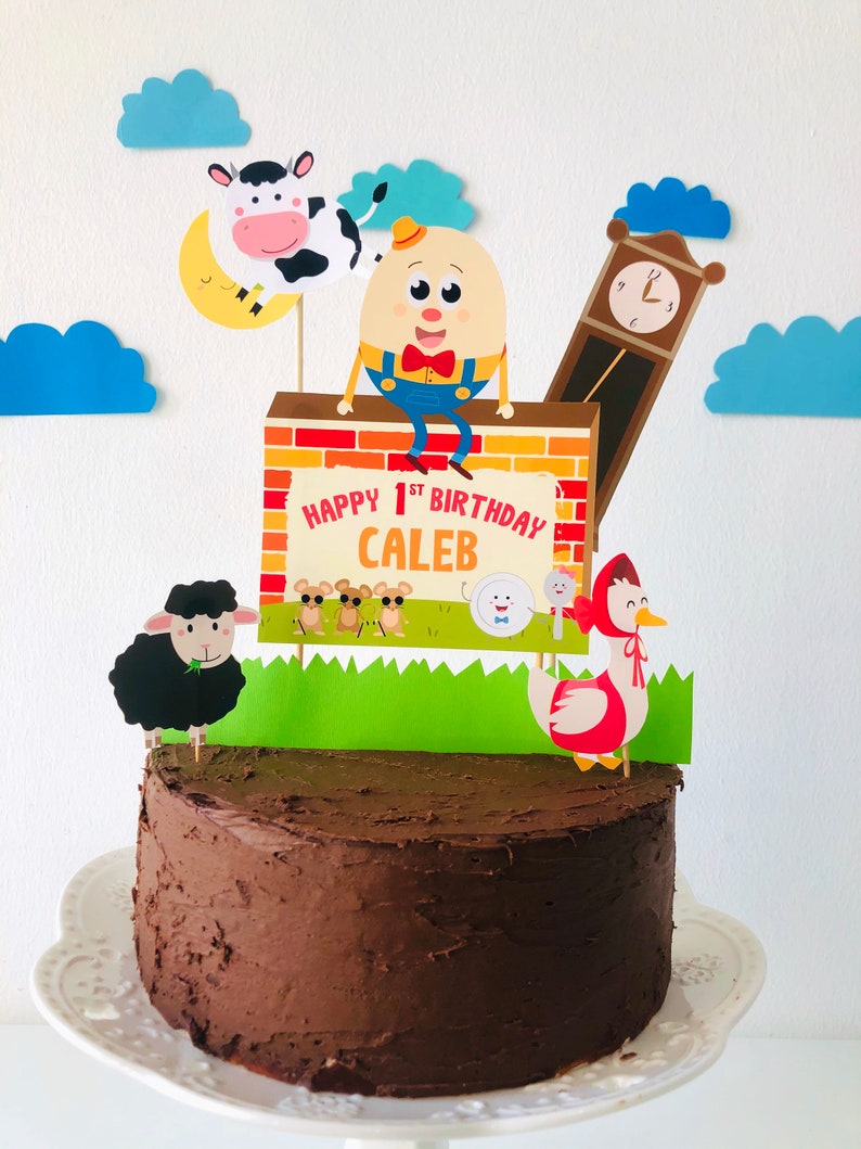 K&ouml;nnte beinhalten: Ein Schokoladenkuchen, der mit einem Banner 'Happy 1st Birthday Caleb', einer Comicfigur eines Eies, einer Kuh, einem Schaf, einer Uhr und einer rotgekappten Gans verziert ist.