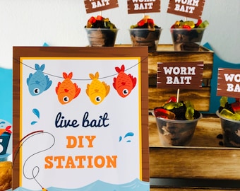May include: A sign with the text "live bait DIY STATION" and a string of four cartoon fish hanging from it. The sign is brown with a yellow border and is set against a wooden background. There are several small cups with chocolate pudding and gummy worms on the table.