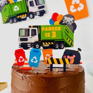 May include: A chocolate cake decorated with a green garbage truck, recycling bins, a construction sign, and a raccoon. The truck has the words "Parker is 3" on the side.