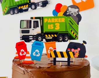 May include: A chocolate cake decorated with a green garbage truck, recycling bins, a construction sign, and a raccoon. The truck has the words "Parker is 3" on the side. 