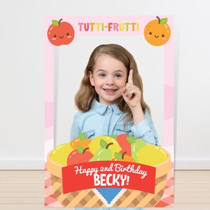 May include: A colorful photo booth prop featuring a basket of fruit with the text "Happy 2nd Birthday Becky!" and the words "Tutti-Frutti" at the top. The prop has a space for a person's face to be placed in the center.