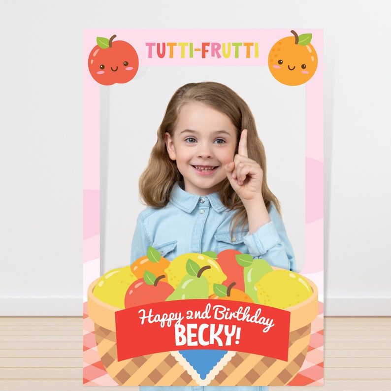 May include: A colorful photo booth prop featuring a basket of fruit with the text "Happy 2nd Birthday Becky!" and the words "Tutti-Frutti" at the top. The prop has a space for a person's face to be placed in the center.