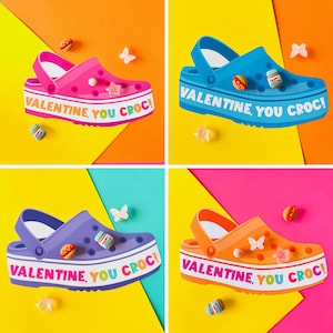 以下が含まれることがあります： 「VALENTINE, YOU CROC!」の文字がソールに書かれた、ピンク、青、紫、オレンジの4色のカラフルなクロックスの靴。チャームで飾られています。背景は黄色、オレンジ、ピンクのミックスです。