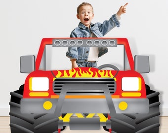 以下が含まれることがあります： 運転席に立っている子供がいる赤いモンスタートラックの段ボール製切り抜き。トラックはボンネットに炎があり、グリルはグレーです。