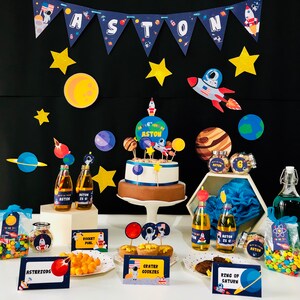 May include: A space-themed birthday party setup with a cake, decorations, and treats. The cake is decorated with planets and a rocket ship. The banner spells out "ASTON". Other decorations include star cutouts, planets, and rocket ships. Treats include "Asteroids" and "Crater Cookies".