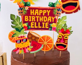 Könnte beinhalten: Ein Schokoladenkuchen, der mit einem tropischen Luau-Thema dekoriert ist. Der Kuchen hat ein Schild mit der Aufschrift "Happy Birthday Ellie" und ist mit Ananas, Wassermelone, Orangenscheiben und einer Tiki-Maske verziert.