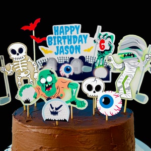 May include: A chocolate cake decorated with a variety of Halloween-themed cake toppers, including a skeleton, a zombie, a mummy, and a golf ball. The cake is decorated with the words "Happy Birthday Jason" and the number "6".