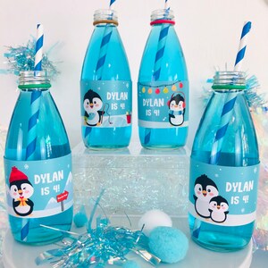 May include: Four blue glass bottles with blue and white striped straws. Each bottle has a personalized label with a penguin design and the text "DYLAN IS 4!"