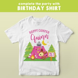 May include: White t-shirt with a pink tent, a raccoon, a deer, and a campfire. The text "Happy Camper Quinn" is above the tent. The number "6" is on a sign next to the deer.