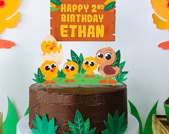 May include: A chocolate cake decorated with a wooden sign that says "Happy 2nd Birthday Ethan" and a variety of yellow and brown chicks. The cake is on a white cake stand and is surrounded by green leaves.