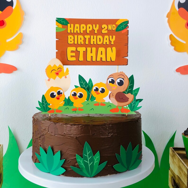 May include: A chocolate cake decorated with a wooden sign that says "Happy 2nd Birthday Ethan" and a variety of yellow and brown chicks. The cake is on a white cake stand and is surrounded by green leaves.