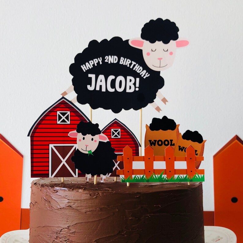 May include: A chocolate cake decorated with a farm theme. The cake has a red barn, a black sheep, a fence, and a sign that says "Happy 2nd Birthday Jacob!"