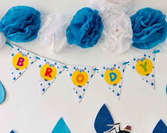 May include: A blue and yellow banner with the word "BRODY" spelled out in white letters. The banner is decorated with small, colorful raindrops and spiders. The banner is hanging from a white wall with blue and white tissue paper decorations.