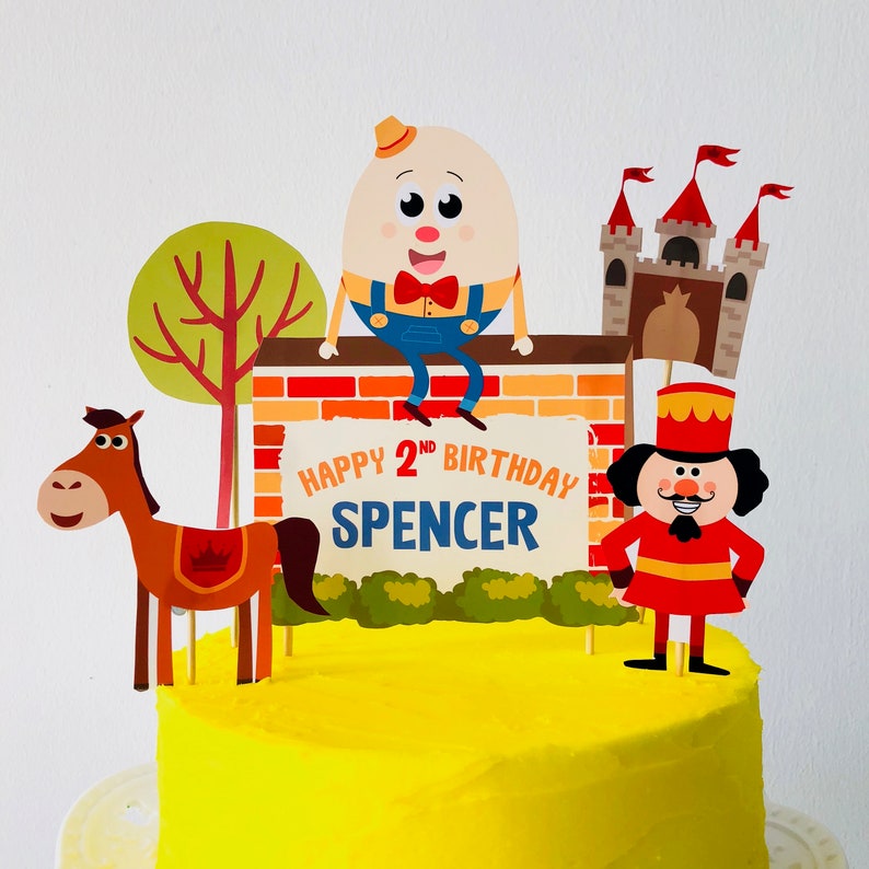 Puede incluir: Un pastel amarillo decorado con una pared de ladrillo, un &aacute;rbol verde, un caballo, un hombre de jengibre y un personaje de huevo. El pastel est&aacute; decorado con el texto "Happy 2nd Birthday Spencer".