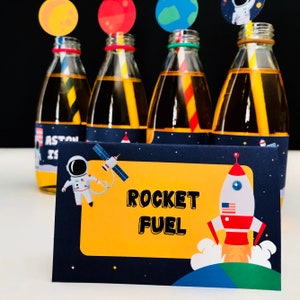 May include: Four glass bottles filled with a yellow liquid, each with a colorful straw and a planet-themed topper. A navy blue card with the words "ROCKET FUEL" and a rocket ship illustration is in front of the bottles. The theme is space and astronomy.