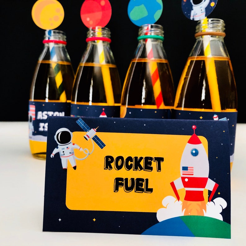 May include: Four glass bottles filled with a yellow liquid, each with a colorful straw and a planet-themed topper. A navy blue card with the words "ROCKET FUEL" and a rocket ship illustration is in front of the bottles. The theme is space and astronomy.