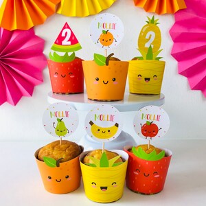 Six decorated cupcake cases with fruit designs. The cupcake wrappers are pineapple, watermelon and orange designs. Each wrapper has a cute cartoon face with eyes and a smile. The cupcakes are topped with cupcake toppers featuring a watermelon slice, a pineapple, a pear, a banana, an apple, and an orange. Each topper has a cartoon face and the name "Mollie" written on it.