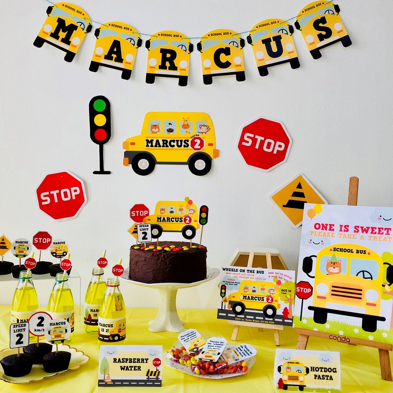 A birthday party table setting with a yellow school bus theme. The table is decorated with yellow school buses, stop signs, and a chocolate cake with a school bus topper. There are also cupcakes, sweets, and drinks on the table. The table is covered in a yellow tablecloth. The text on the cake says "Marcus 2".