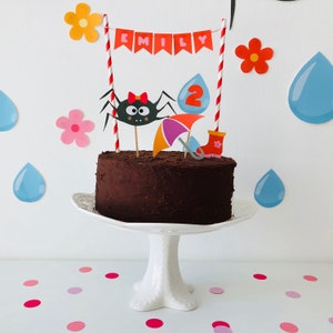 May include: A chocolate cake with a spider, umbrella, rain boots, and the name "Emily" on top. The cake is decorated with colorful paper decorations and is on a white cake stand.
