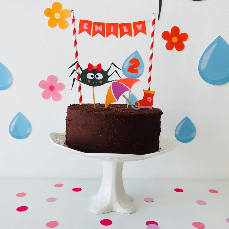 May include: A chocolate cake with a spider, umbrella, rain boots, and the name "Emily" on top. The cake is decorated with colorful paper decorations and is on a white cake stand.