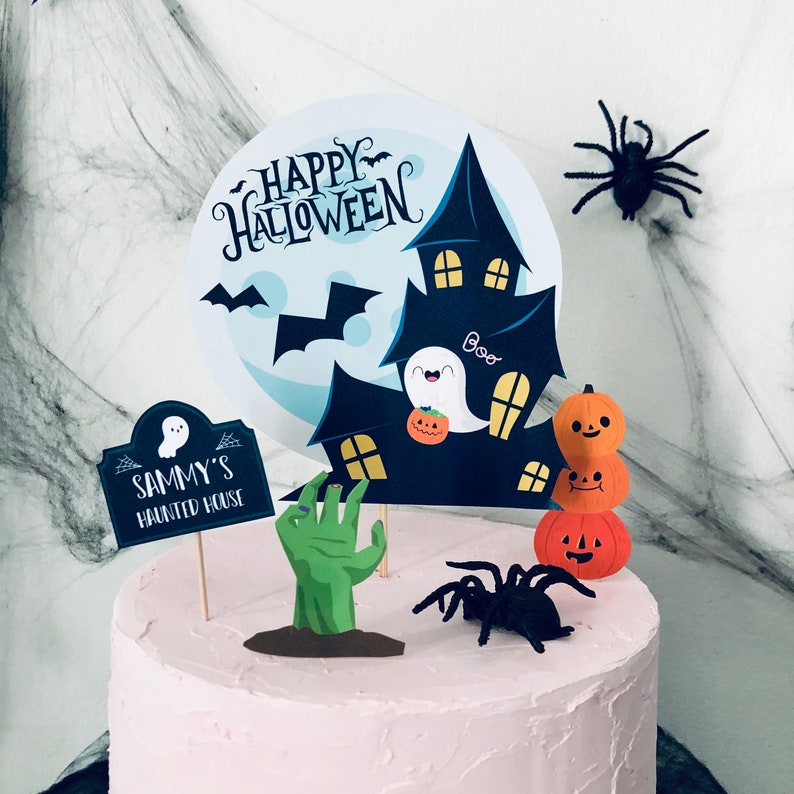 May include: A pink frosted cake decorated with Halloween themed cake toppers. The toppers include a haunted house, a ghost, a zombie hand, a spider, and a stack of pumpkins. The cake is decorated with the words "Happy Halloween" and "Sammy's Haunted House".