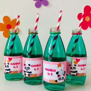 Op de afbeelding: Vier groene glazen flessen met roze en witte etiketten met een pandabeerontwerp. Elke fles heeft een rood-wit gestreepte rietje en de tekst "Hanna is 3".