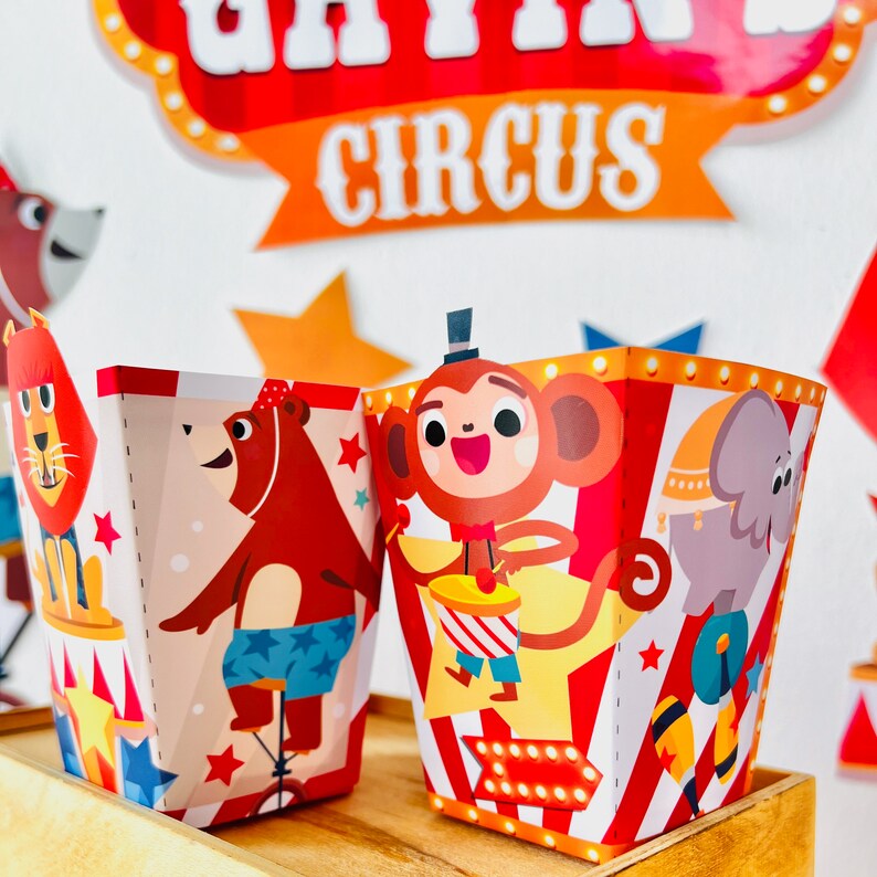May include: Two colorful party favor boxes with circus animal designs. The box on the left features a brown bear wearing a blue and white striped swimsuit riding a unicycle. The box on the right features a monkey wearing a red and white striped outfit playing a drum. Both boxes have a red and white striped background with yellow stars.