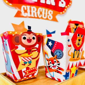 May include: Two colorful party favor boxes with circus animal designs. The box on the left features a monkey playing the drums, while the box on the right features a tiger jumping through a hoop. Both boxes have red and white stripes and yellow stars.