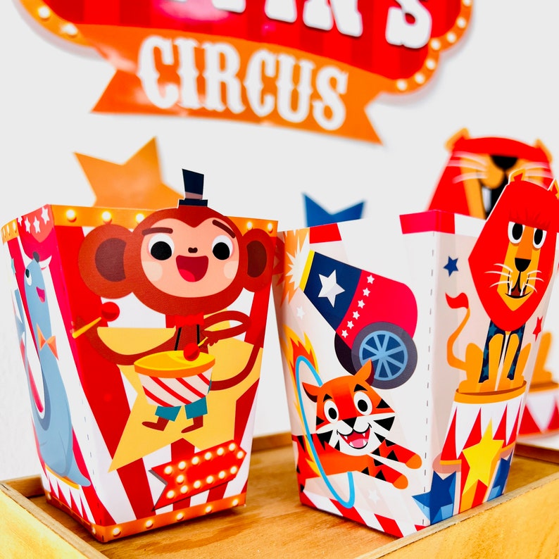 May include: Two colorful party favor boxes with circus animal designs. The box on the left features a monkey playing the drums, while the box on the right features a tiger jumping through a hoop. Both boxes have red and white stripes and yellow stars.