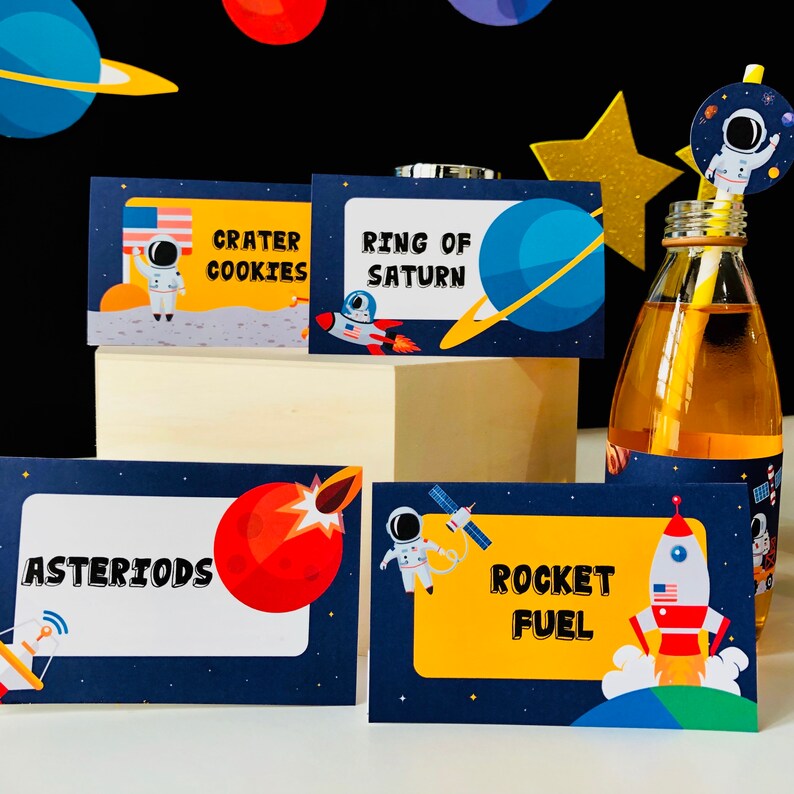 May include: A collection of space-themed party decorations. Includes navy blue and yellow cards with illustrations of planets, rockets, and astronauts. Text on the cards reads "Crater Cookies", "Ring of Saturn", "Asteroids", and "Rocket Fuel". A bottle with a straw and space-themed decorations is also present.