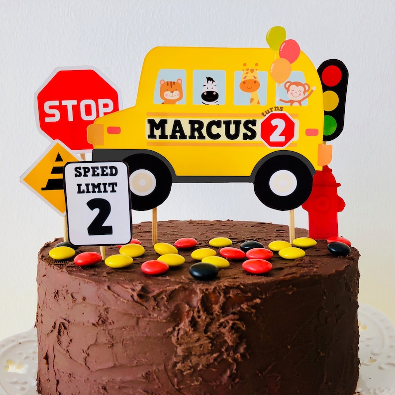 A Wheels on the Bus themed birthday cake decorated with a yellow school bus cake topper, red and yellow M&Ms, and a speed limit sign with the number 2. The bus has the name "MARCUS" and the number "2" on it.