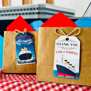 May include: Two brown paper gift bags with blue and red tissue paper peeking out. Each bag has a white tag with a blue and white illustration of a ship and the text "Thank you for coming. Love, Johnny."
